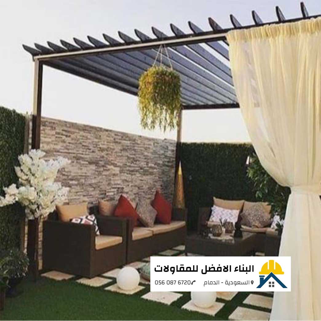 الرئيسية - البناء الافضل للمقاولات برجولة جلسة حدائق حديد شكل خشب ذات سقف مفرغ ومزينة بستائر بيضاء ناعمة ونباتات معلقة، تحتوي على طقم كنب خارجي بني، جدار حجر تشديد، وأرضية عشب صناعي مع بلاط، من تنفيذ مؤسسة البناء الأفضل للمقاولات بالدمام مع الشعار ورقم الاتصال 0560876720.