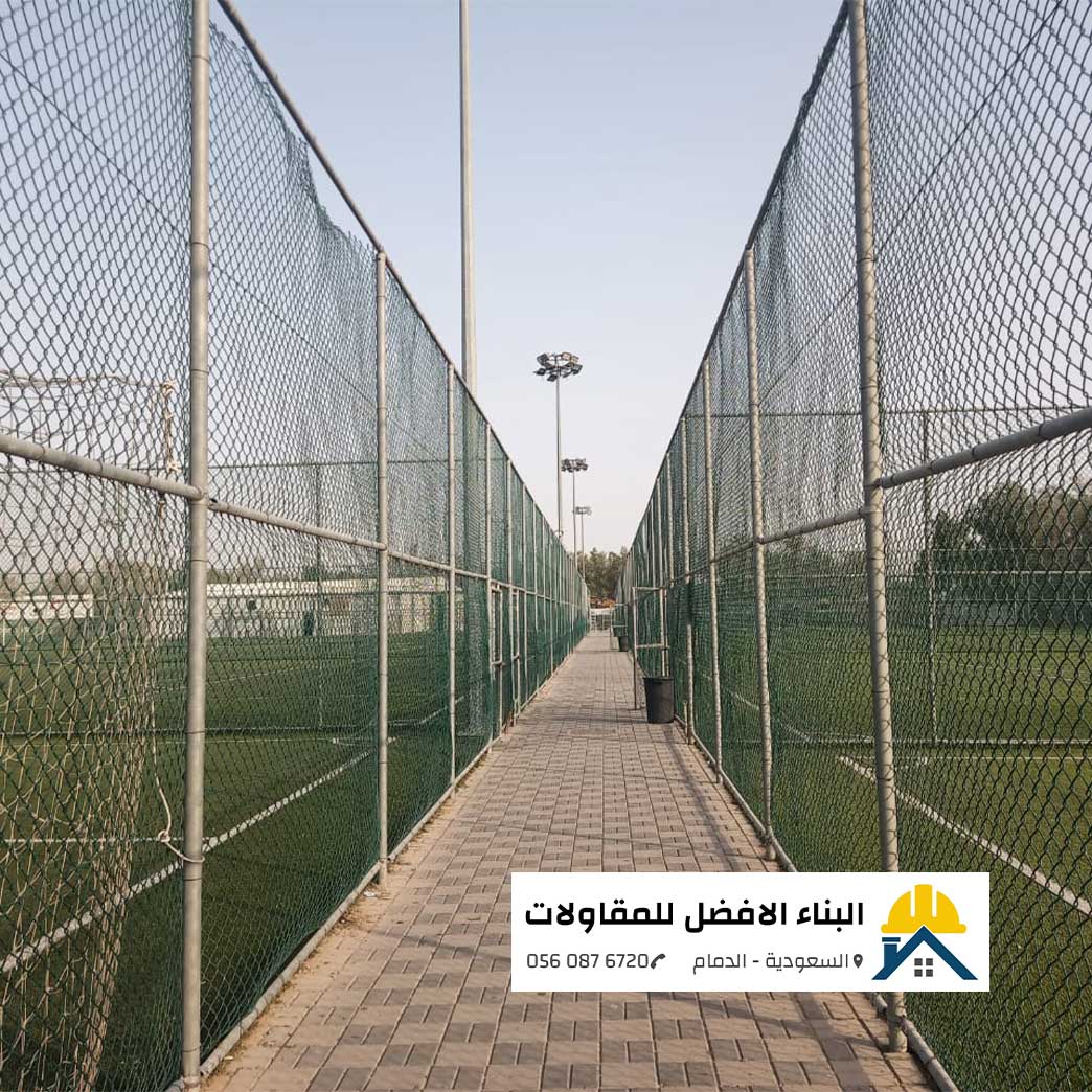 الرئيسية - البناء الافضل للمقاولات ممر مرصوف بالبلاط المتداخل (الإنترلوك) يفصل بين ملعبين رياضيين، محاط بشباك وشباك شبكية مجلفنة عالية الارتفاع مثبتة على أنابيب حديدية قوية لحماية الملاعب، مع وجود كشافات إضاءة وشعار مؤسسة البناء الأفضل للمقاولات بالدمام ورقم التواصل 0560876720 في الأسفل.