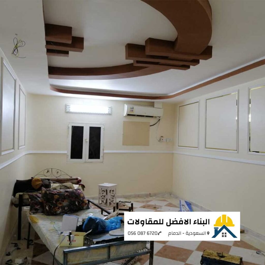 غرفة منزلية مدهونة بأصباغ داخلية ناعمة باللون البيج الفاتح تحتوي على إطارات ديكورات فوم جدارية مع خطوط استيل ذهبي وسقف ديكوري من الجبس بورد بألوان بني وبيج، من تنفيذ مؤسسة البناء الأفضل للمقاولات بالدمام مع الشعار ورقم الاتصال 0560876720.
