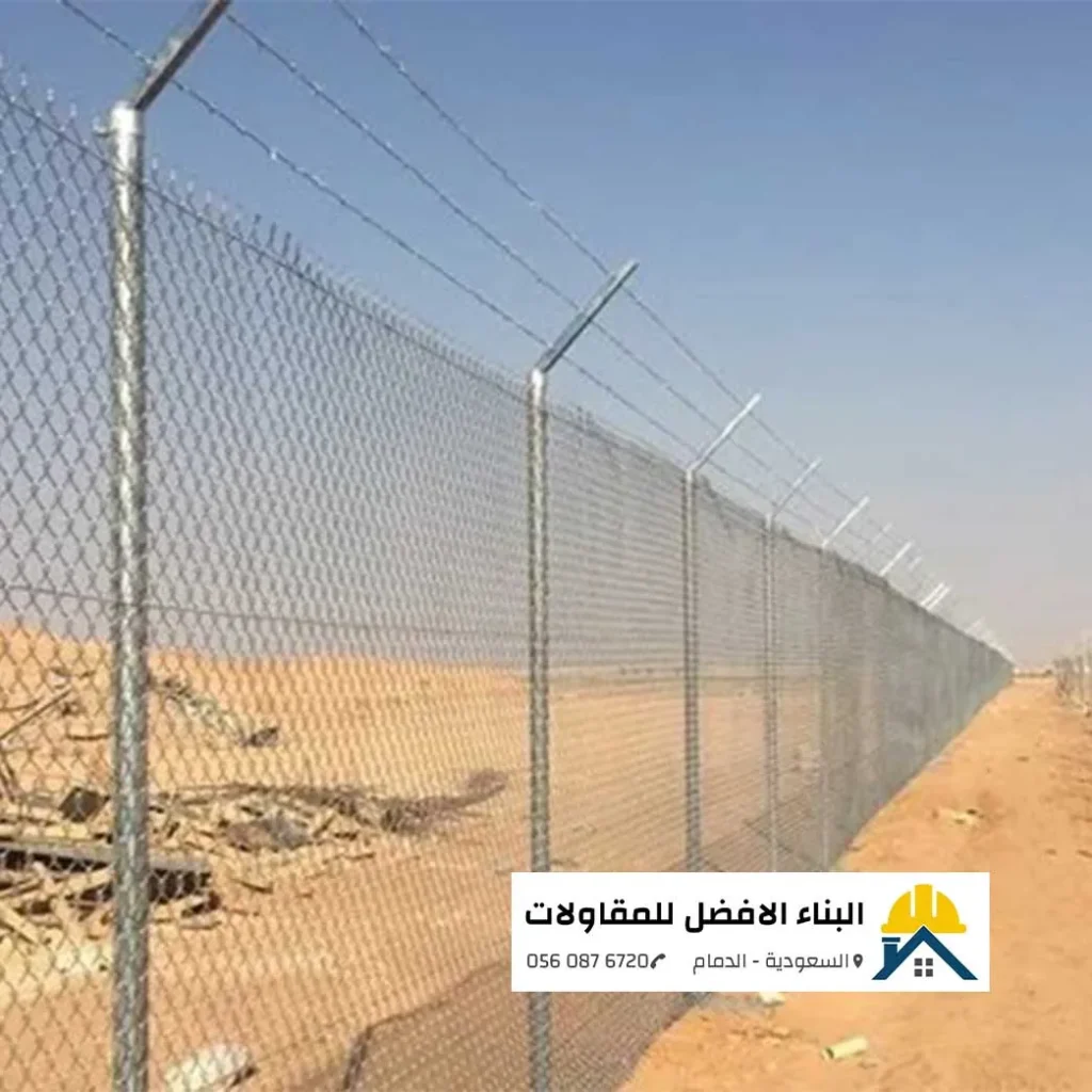 سياج شبكي مجلفن مرتفع وممتد على مسافة طويلة في أرض صحراوية بالدمام، مزود بأسلاك شائكة علوية مشدودة بدقة لحماية المنشآت، مع شعار البناء الأفضل للمقاولات.