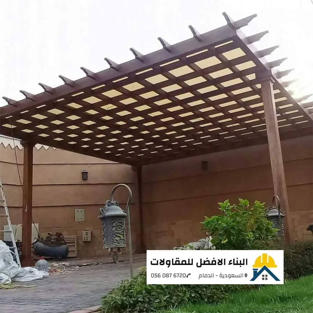 هيكل برجولة خشبية ضخمة بنظام المربعات والمجدول مع طبقة تغطية عازلة تحت السقف، مثبتة في حوش فيلا بجانب جدار خارجي بتصميم تراثي وفوانيس أرضية كلاسيكية، مع ظهور معدات التركيب والسلم في موقع العمل، من تنفيذ مؤسسة البناء الأفضل للمقاولات بالدمام 0560876720.