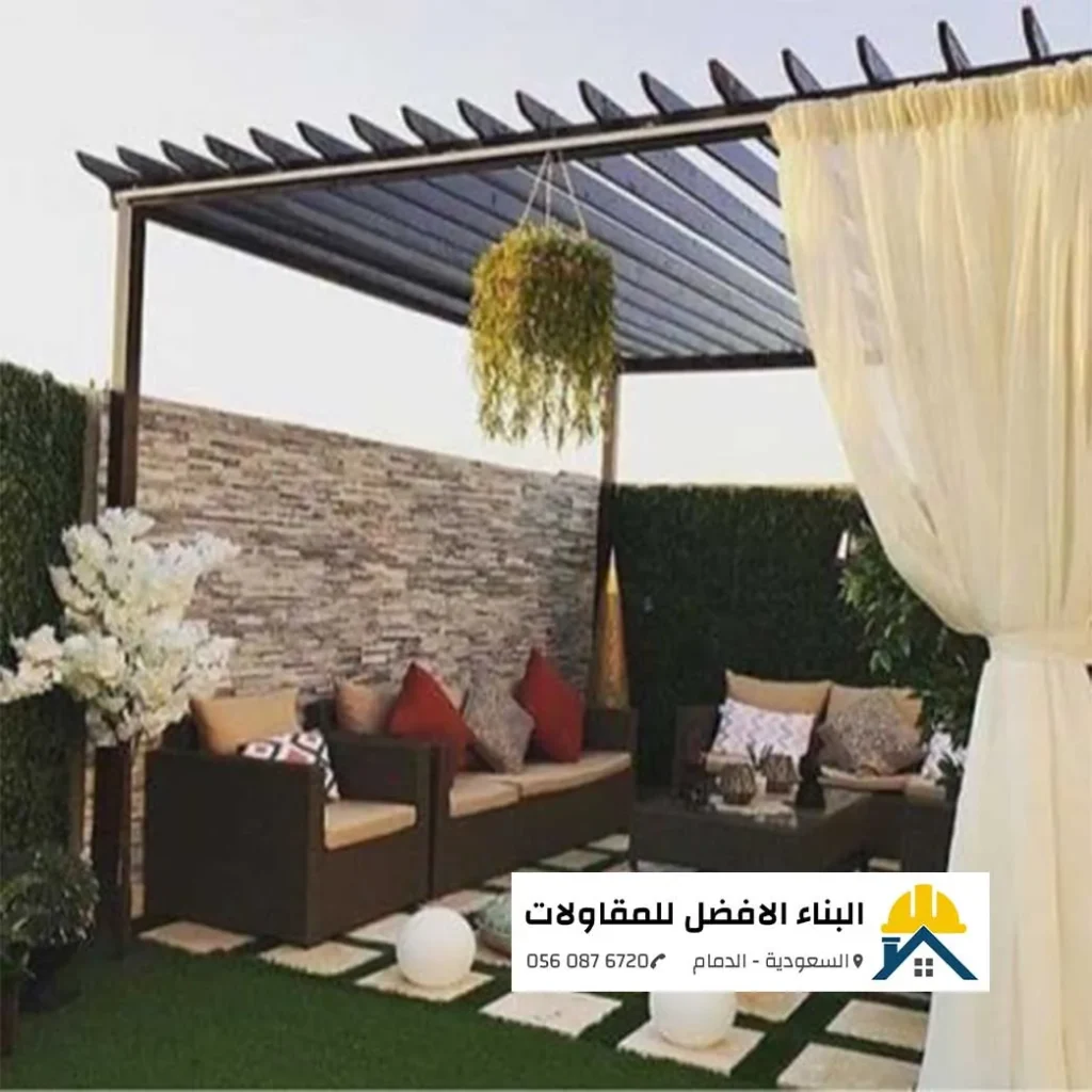 برجولة مودرن حديد شكل خشب بلون بني غامق مزينة بنباتات خضراء معلقة وستائر بيضاء جانبية ناعمة، وتحتها جلسة كنب حدائق راتان بلون بني ووسائد حمراء فوق أرضية ممر مربعات وعشب صناعي، من تنفيذ مؤسسة البناء الأفضل للمقاولات بالدمام 0560876720.
