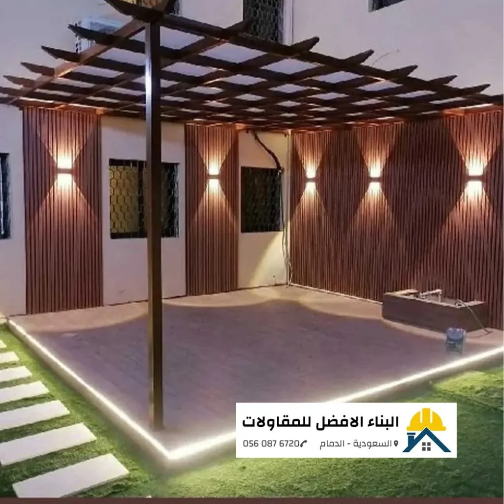 جلسة حدائق خارجية فاخرة تحت برجولة حديد مودرن بسقف شبكي مدهونة بلون بني خشبى، مع خلفية جدران من التكسيات الخشبية الرأسية المضاءة بإنارة جدارية هندسية وإطار إضاءة ليد أرضي حول المنصة، من تنفيذ مؤسسة البناء الأفضل للمقاولات بالدمام 0560876720.