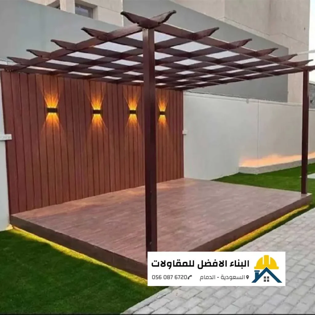 تصميم برجولة حديد مودرن بدهان بني شكل الخشب مع جدار خلفي من التكسيات الخشبية الرأسية المضاءة بإنارة جدارية دافئة، ومنصة أرضية خشبية محاطة بإضاءة ليد مخفية وعشب أخضر، من تنفيذ مؤسسة البناء الأفضل للمقاولات بالدمام 0560876720.