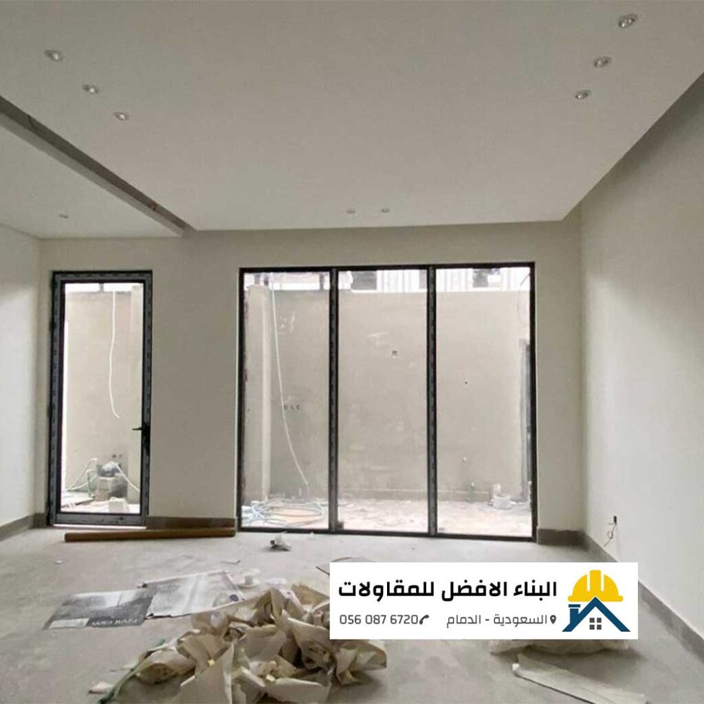 صالة منزلية قيد التشطيب النهائي مدهونة بأصباغ داخلية ناعمة باللون الأوف وايت العصرى، مع أسقف جبس بورد مدمجة بإضاءة سبوت لايت وأبواب زجاجية كبيرة بإطار ألمنيوم أسود، من تنفيذ مؤسسة البناء الأفضل للمقاولات بالدمام مع الشعار ورقم الاتصال 0560876720.