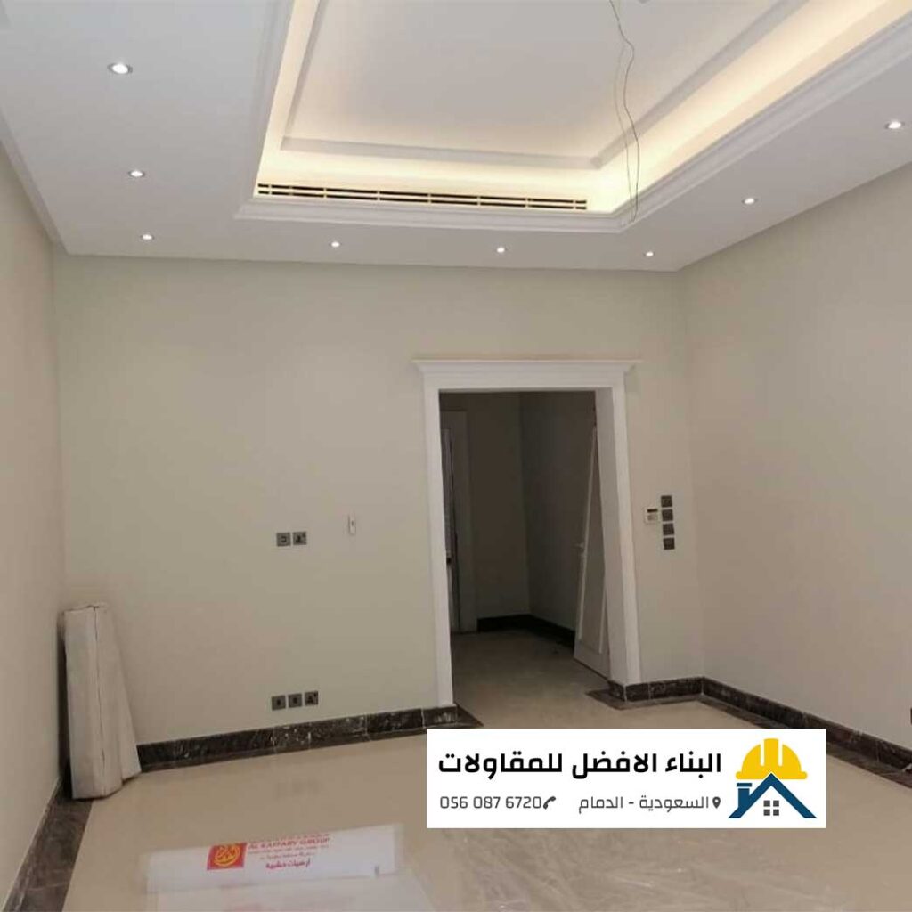 غرفة منزلية مدهونة بأصباغ داخلية ناعمة باللون البيج الفاتح مع سقف جبس بورد مدمج بإضاءة بيت نور وإطار باب خشبي أبيض، من تنفيذ مؤسسة البناء الأفضل للمقاولات بالدمام مع الشعار ورقم الاتصال 0560876720.