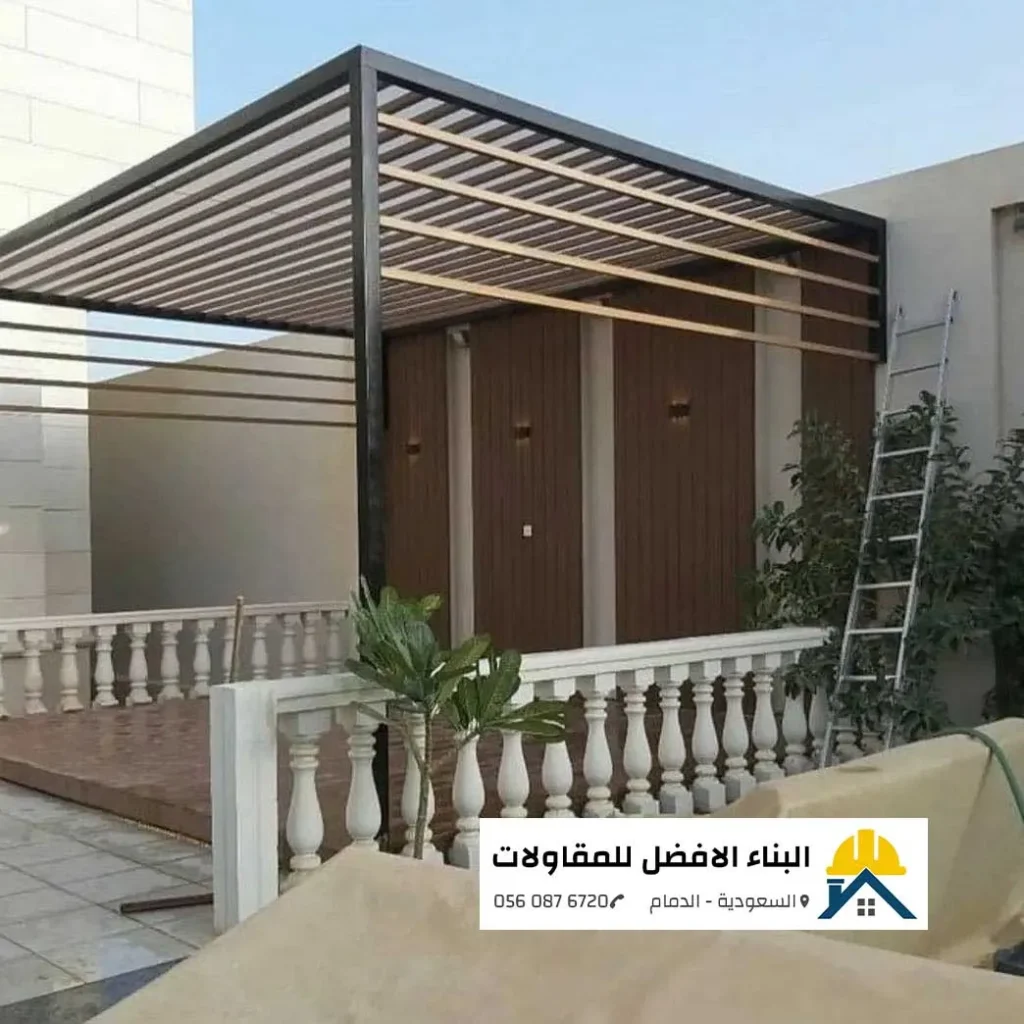 هيكل برجولة حديد مودرن بلون أسود وشرائح أفقية شكل خشب قيد التركيب والتنفيذ الميداني على رووف منزل، مع جدار خلفي مغطى بالتكسيات الخشبية المضاءة بإنارة جدارية وسور بالكونة أبيض، من أعمال مؤسسة البناء الأفضل للمقاولات بالدمام 0560876720.