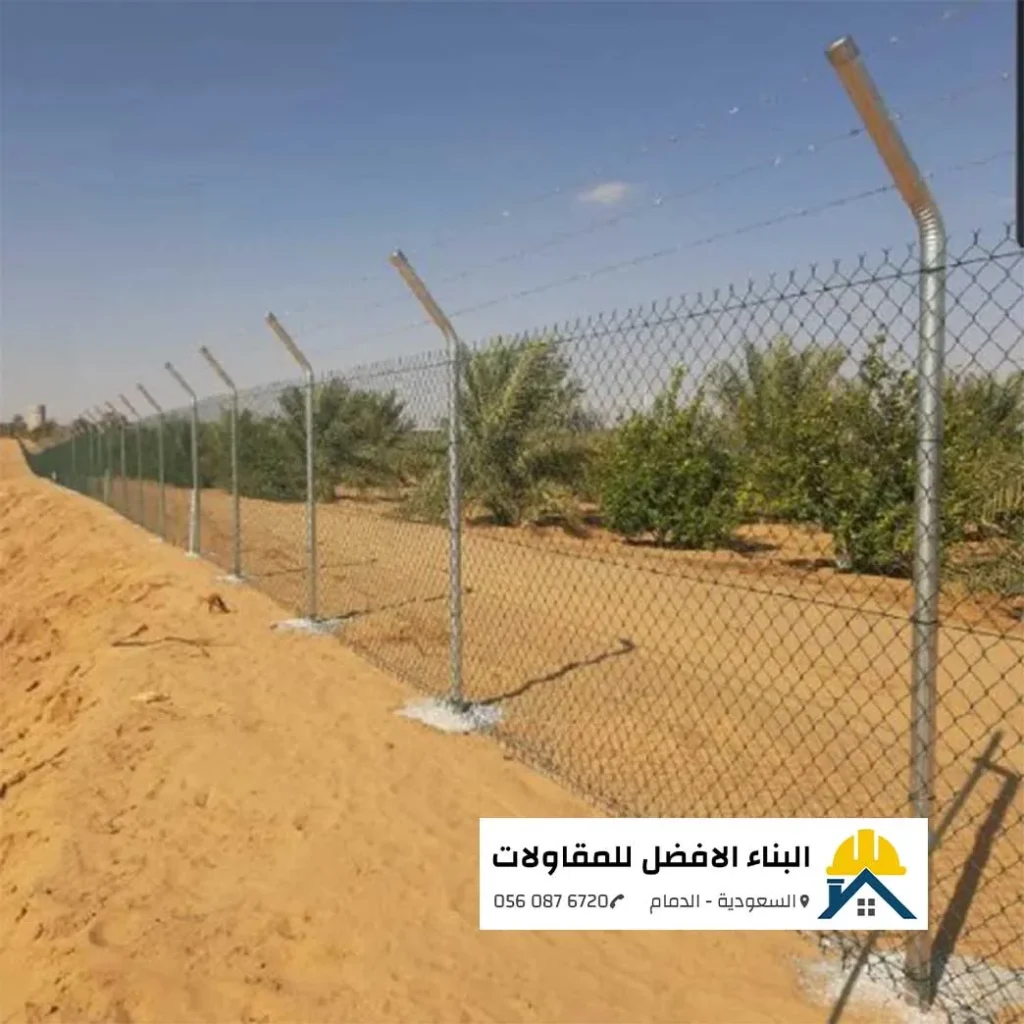صورة لسياج شبكي معدني مجلفن عالي الجودة مركب فوق كثبان رملية مع أسلاك شائكة علوية، تحمي منطقة خضراء (مزرعة) في الدمام. يظهر في زاوية الصورة شعار "البناء الأفضل للمقاولات".