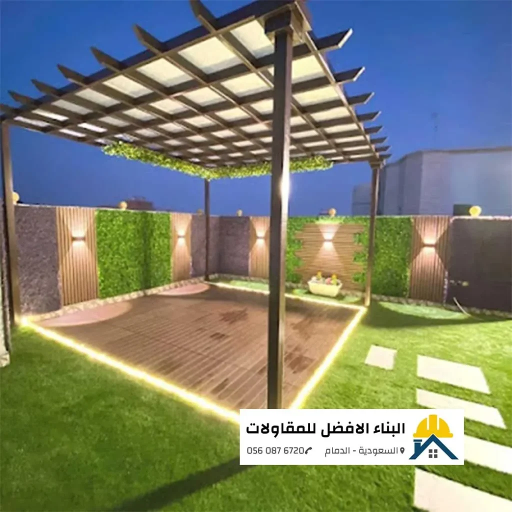 جلسة حدائق خارجية متكاملة تحت برجولة حديد مودرن شكل خشب مزينة بنباتات خضراء متسلقة فوق منصة أرضية خشبية محاطة بشريط إضاءة ليد دافئ، مع جدران من التكسيات الخشبية والعشب الجداري المضاء، من تنفيذ مؤسسة البناء الأفضل للمقاولات بالدمام 0560876720.