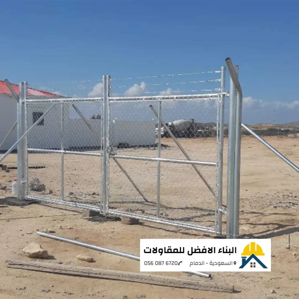 بوابة شبك حديدية مجلفنة مزدوجة الفتح مع أسلاك شائكة علوية مثبتة على أعمدة معدنية سميكة ودعامات مائلة في موقع إنشاء بالدمام، مع شعار البناء الأفضل للمقاولات.