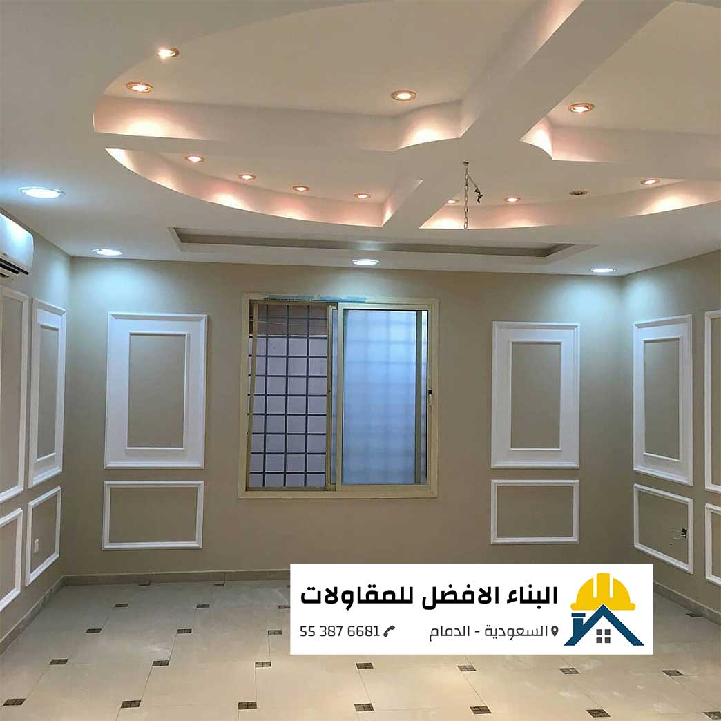 الرئيسية - البناء الافضل للمقاولات تشطيبات وديكورات داخلية لغرفة من تنفيذ مؤسسة البناء الأفضل للمقاولات بالدمام، تظهر فيها دهانات باللون البيج (البيج/الرمادي الهادئ) مع إطارات فوم بيضاء (براقع/فوم جداري) على الجدران، وسقف جبس بورد ديكوري مزين بإضاءات مخفية وسبوت لايت، يتوسط الغرفة نافذة بألمنيوم زجاجي، وفي الأسفل يظهر شعار المؤسسة مع رقم الاتصال.